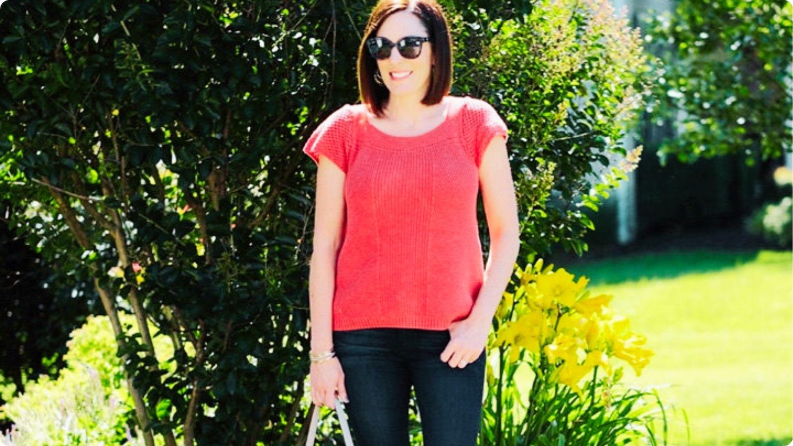Woman stands in a garden wearing a coral-pink knit top and dark blue skinny jeans.
