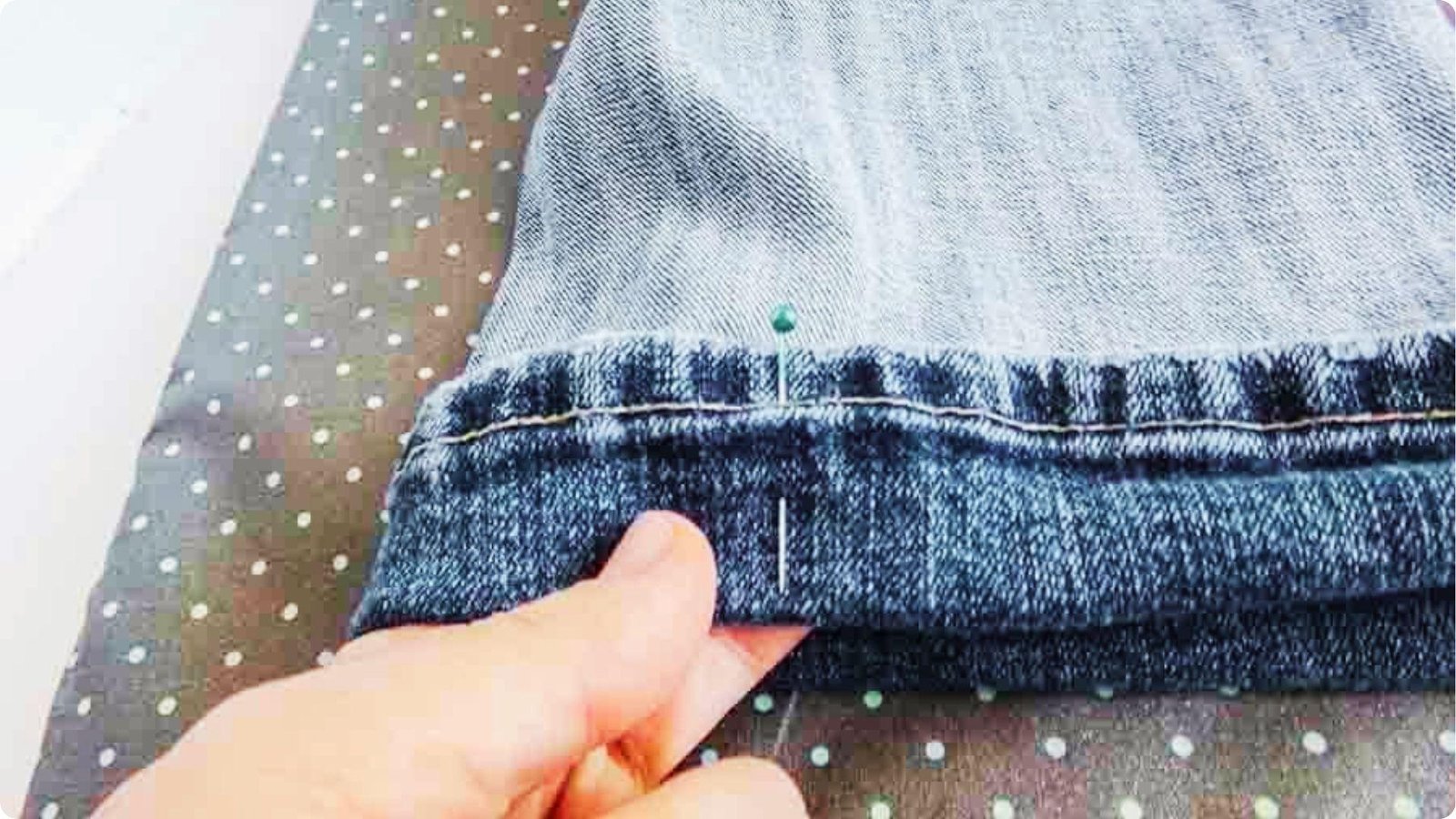 A hand pins the folded hem of a denim pant leg with a green sewing pin on a polka-dot ironing board.