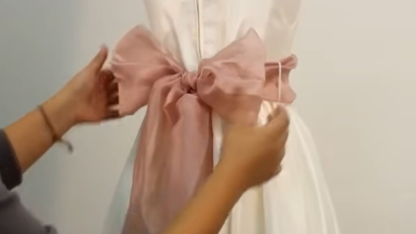 "Hands adjusting a large dusty pink satin bow tied at the back of a white dress with a zipper closure"