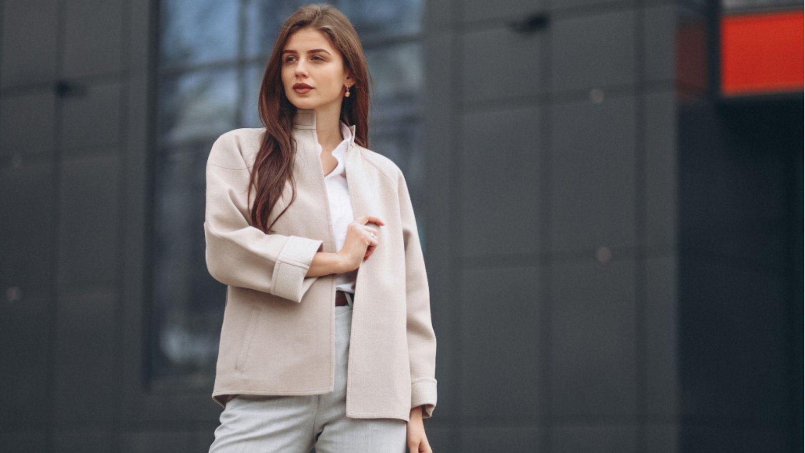 A brunette woman posing outdoors in a beige collarless coat, white shirt, and grey trousers against a dark modern building.