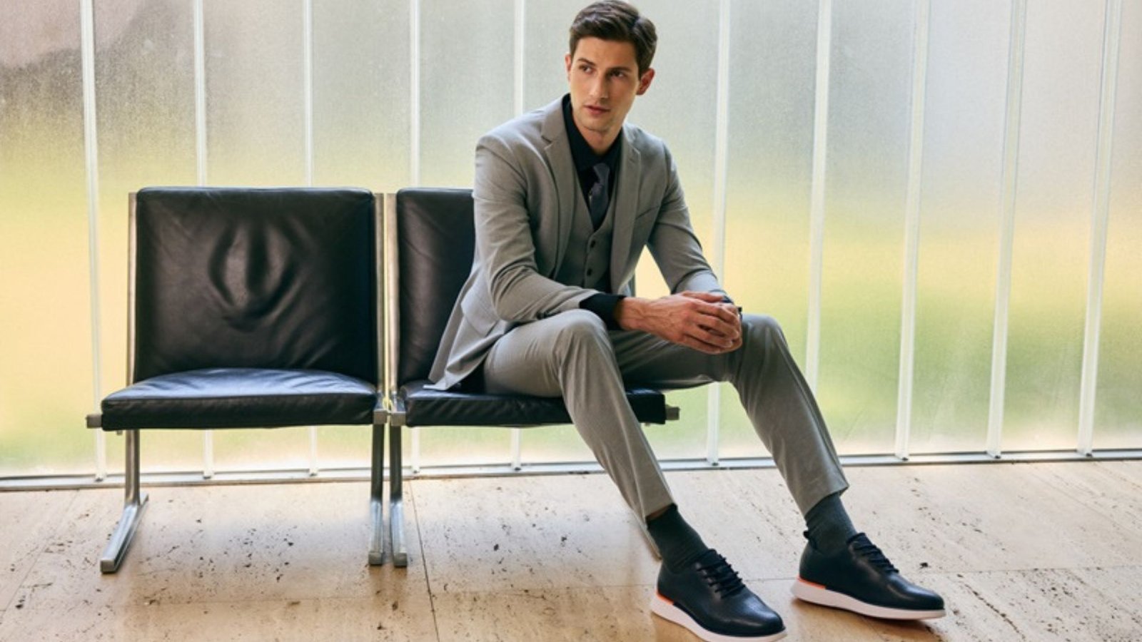 Man wearing grey dress pants and blazer with navy blue sneakers, sitting on a black leather chair in a modern indoor setting