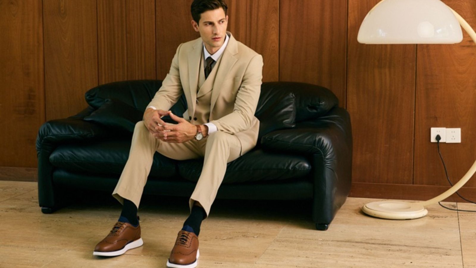 Man wearing beige dress pants and blazer with brown oxford sneakers, sitting on a black leather sofa in a stylish indoor setting
