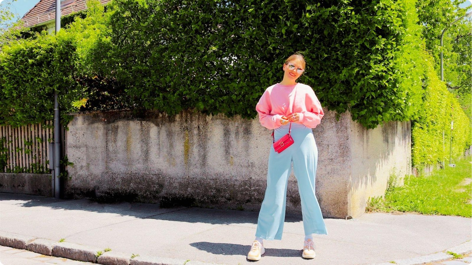 Woman smiles on a sunny street in a baby pink sweatshirt, powder blue wide-leg trousers, and pink crossbody bag.