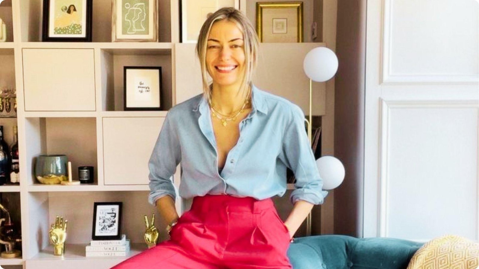Woman sits indoors wearing a light blue button-down shirt tucked into bright pink trousers.