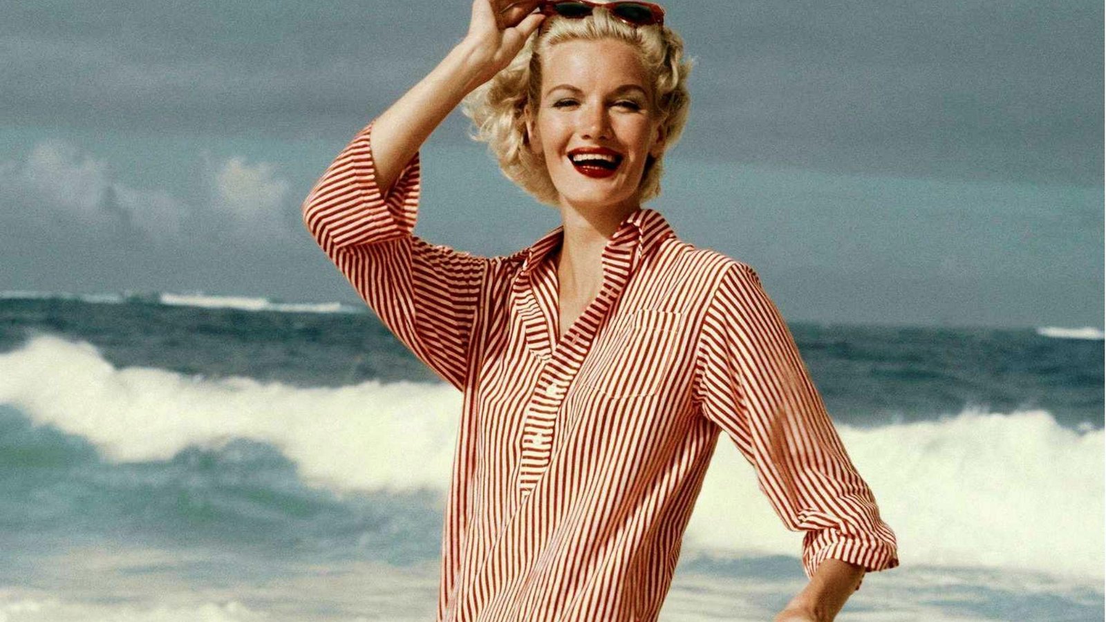 A vintage photograph of a woman standing on a beach in flowy red and white linen shirt, depicting coastal grandmother style.