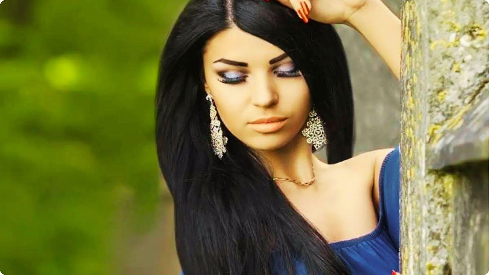 Woman with long black hair wears large silver chandelier earrings and a gold necklace with a navy blue off-shoulder dress.