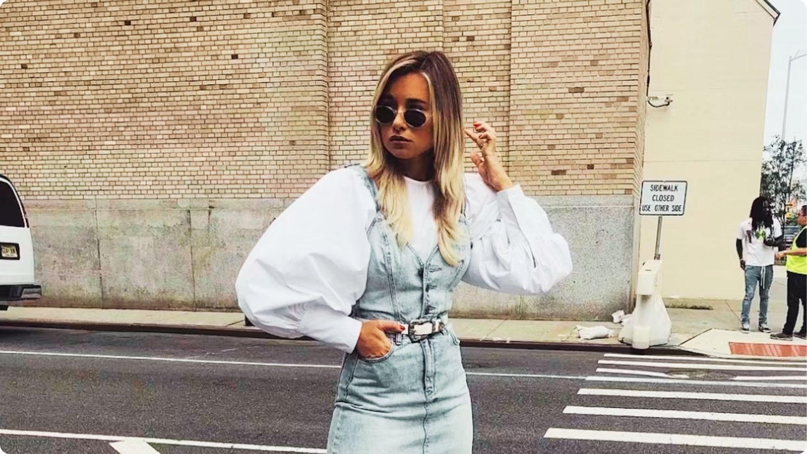 Woman posing at a crosswalk in a white balloon-sleeve shirt layered under a light-wash denim pinafore dress.