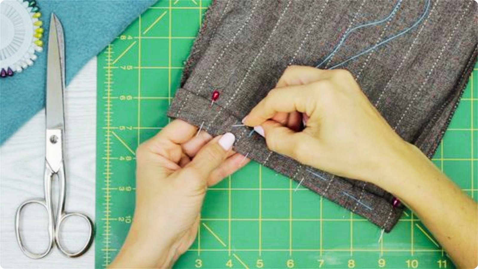 Hands pinning the hem of gray pinstripe dress pants on a green cutting mat, with scissors and pins nearby.