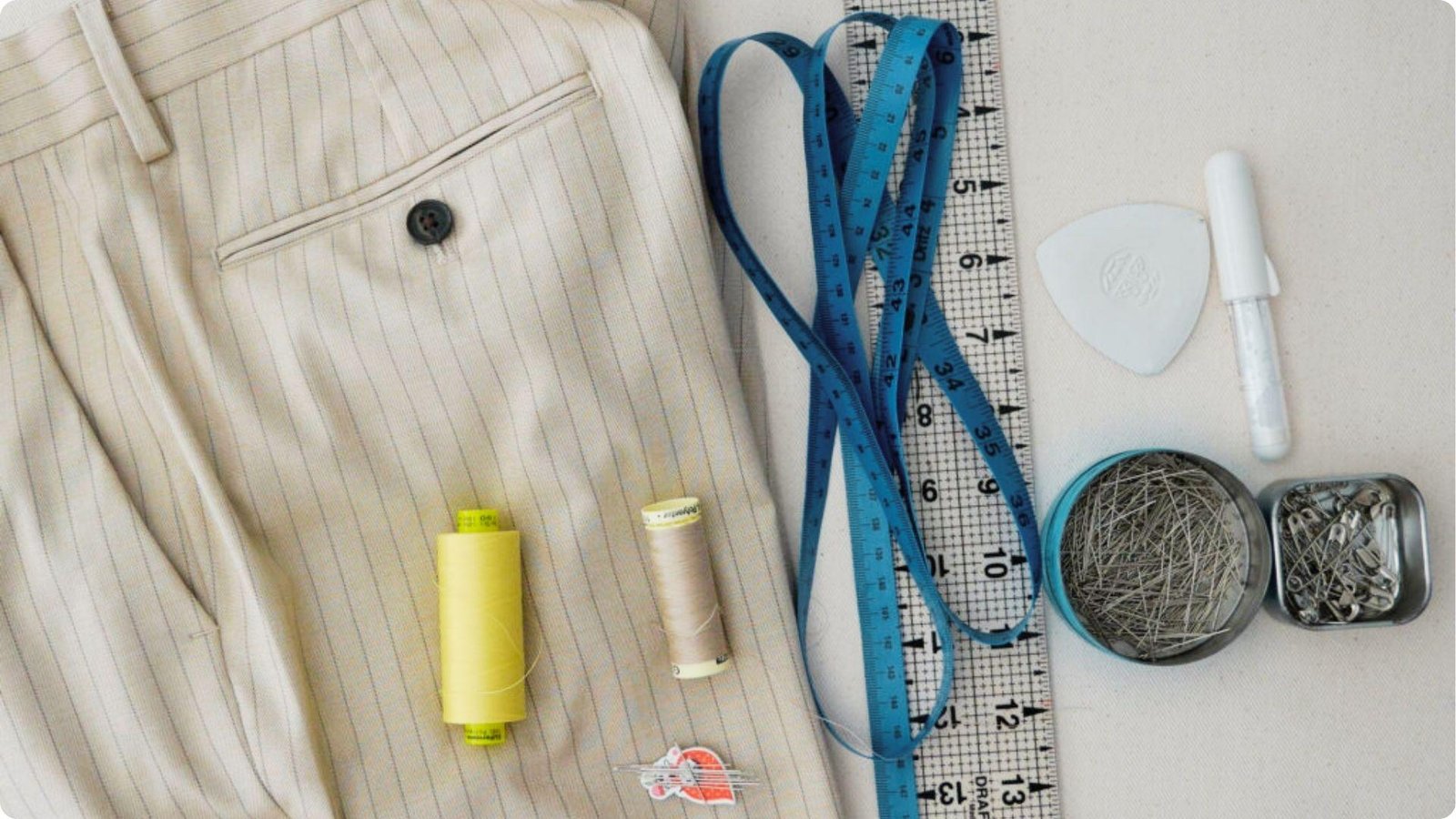 Flat lay of hemming supplies on a table: beige pinstripe dress pants, measuring tape, ruler, pins, and thread spools.
