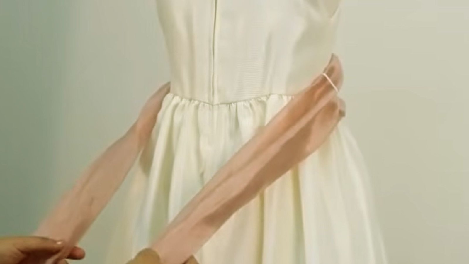 "Hands holding both ends of a dusty pink satin sash wrapped around the waist of a white dress, preparing to tie it"