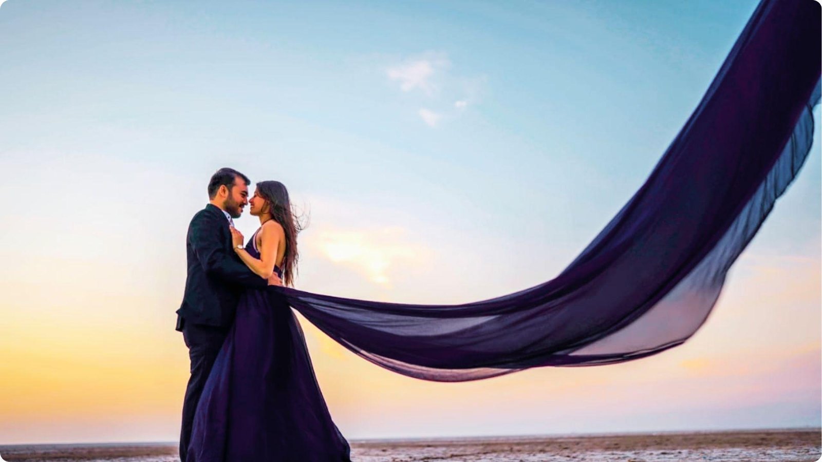 Couple embraces at sunset as the woman’s flowing navy blue gown with sheer train blows in the wind.