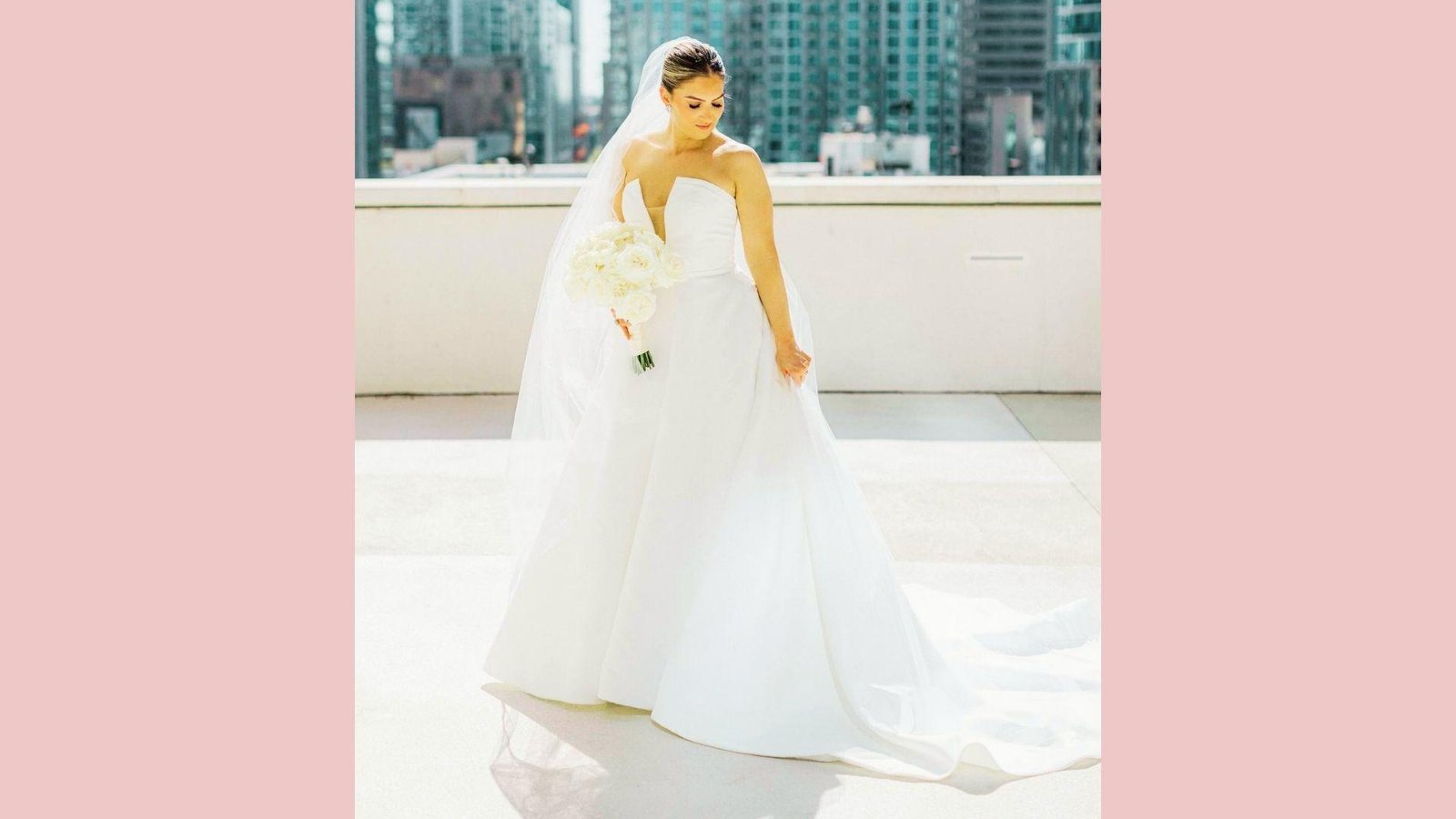 Bride on rooftop wearing strapless A-line satin gown with plunging neckline, long veil, holding bouquet, city skyline behind