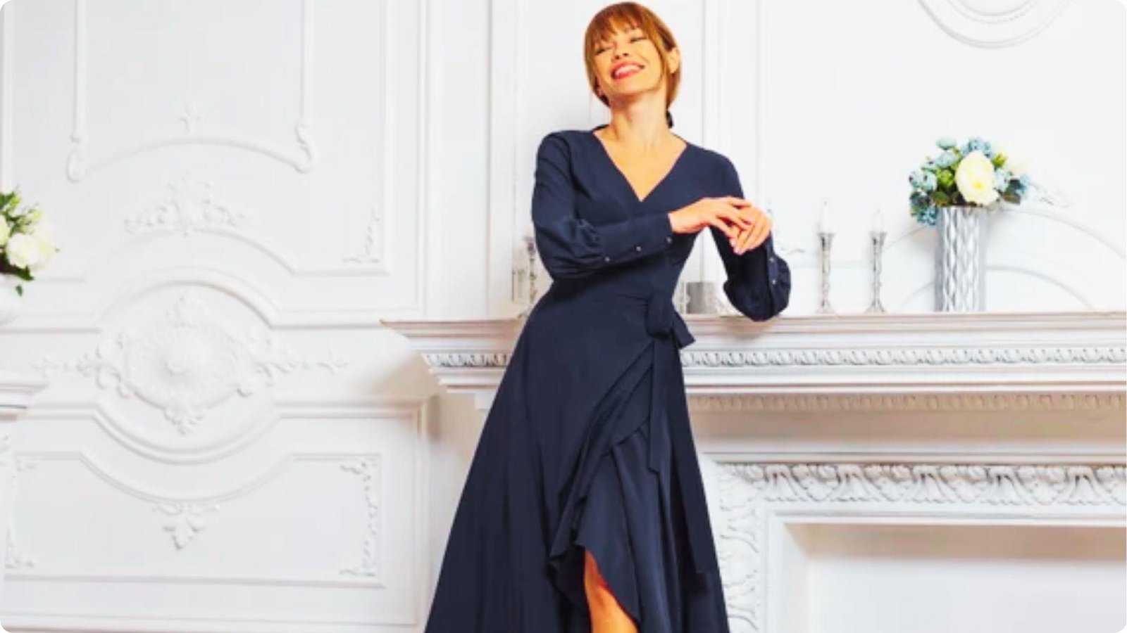 Smiling woman in a navy blue wrap dress with long sleeves and a high-low hem poses in a white formal room.