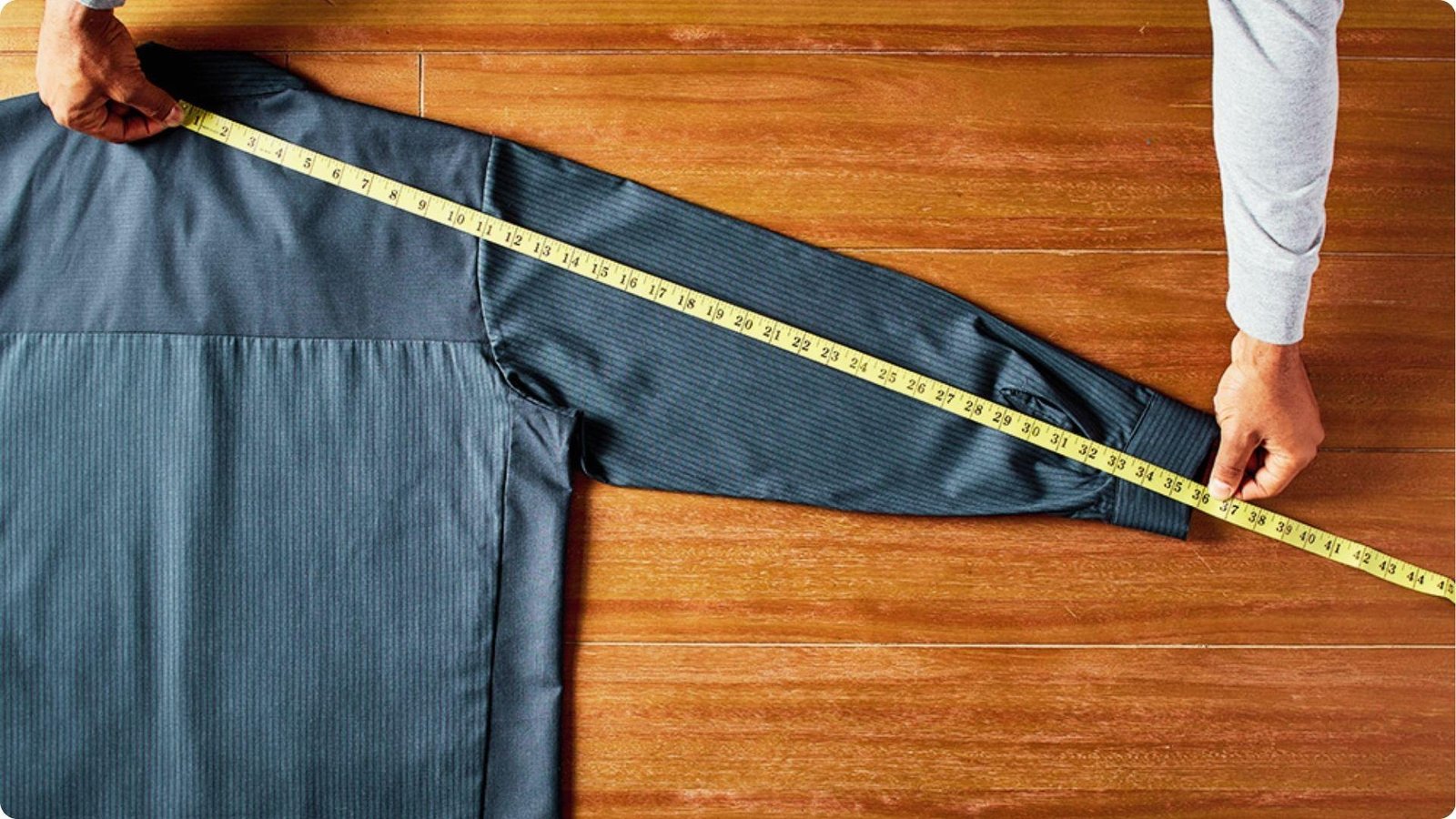 Hands holding a yellow measuring tape across a dark dress shirt laid on a wood floor, measuring from center back collar to cuff.