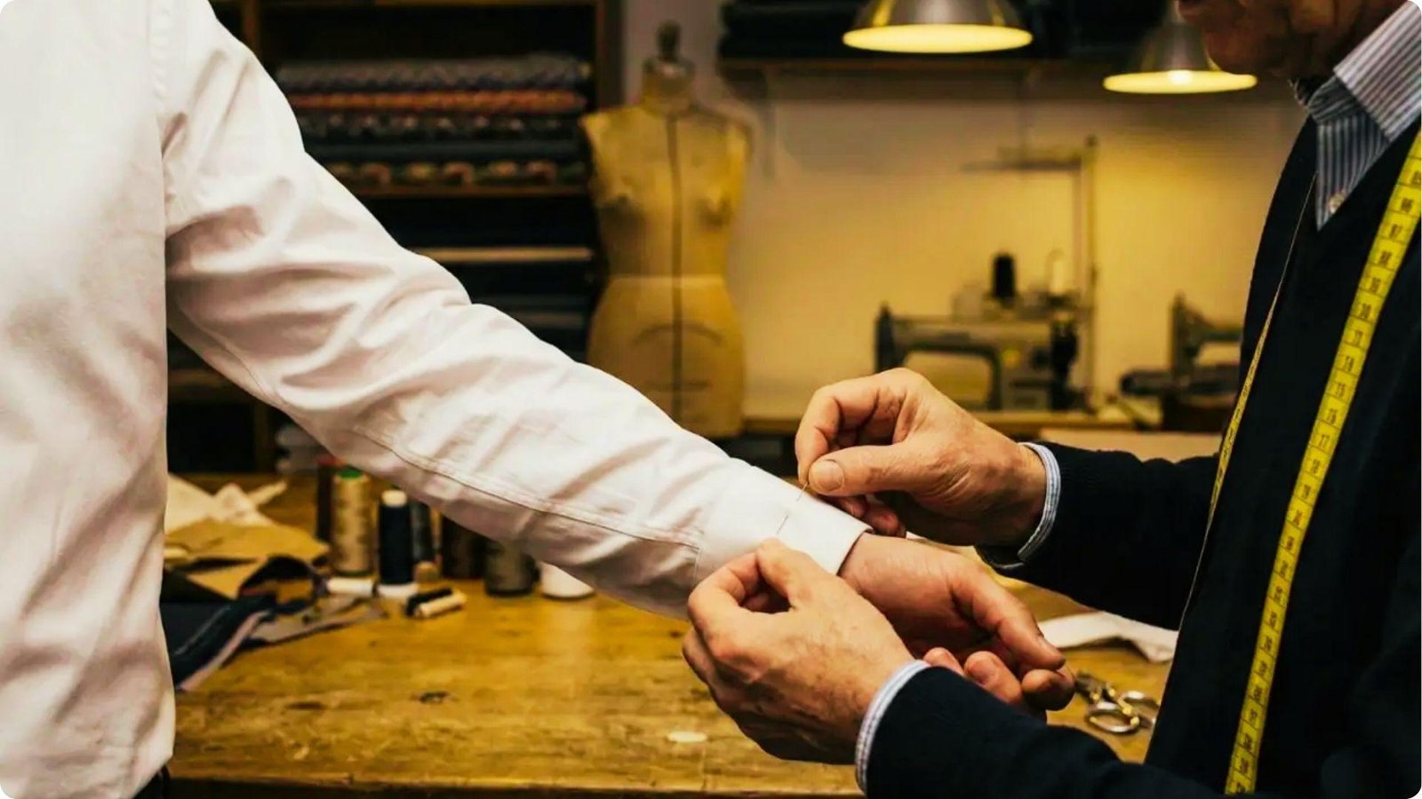 Tailor pinning a dress shirt cuff in a workshop with measuring tape draped around his neck.