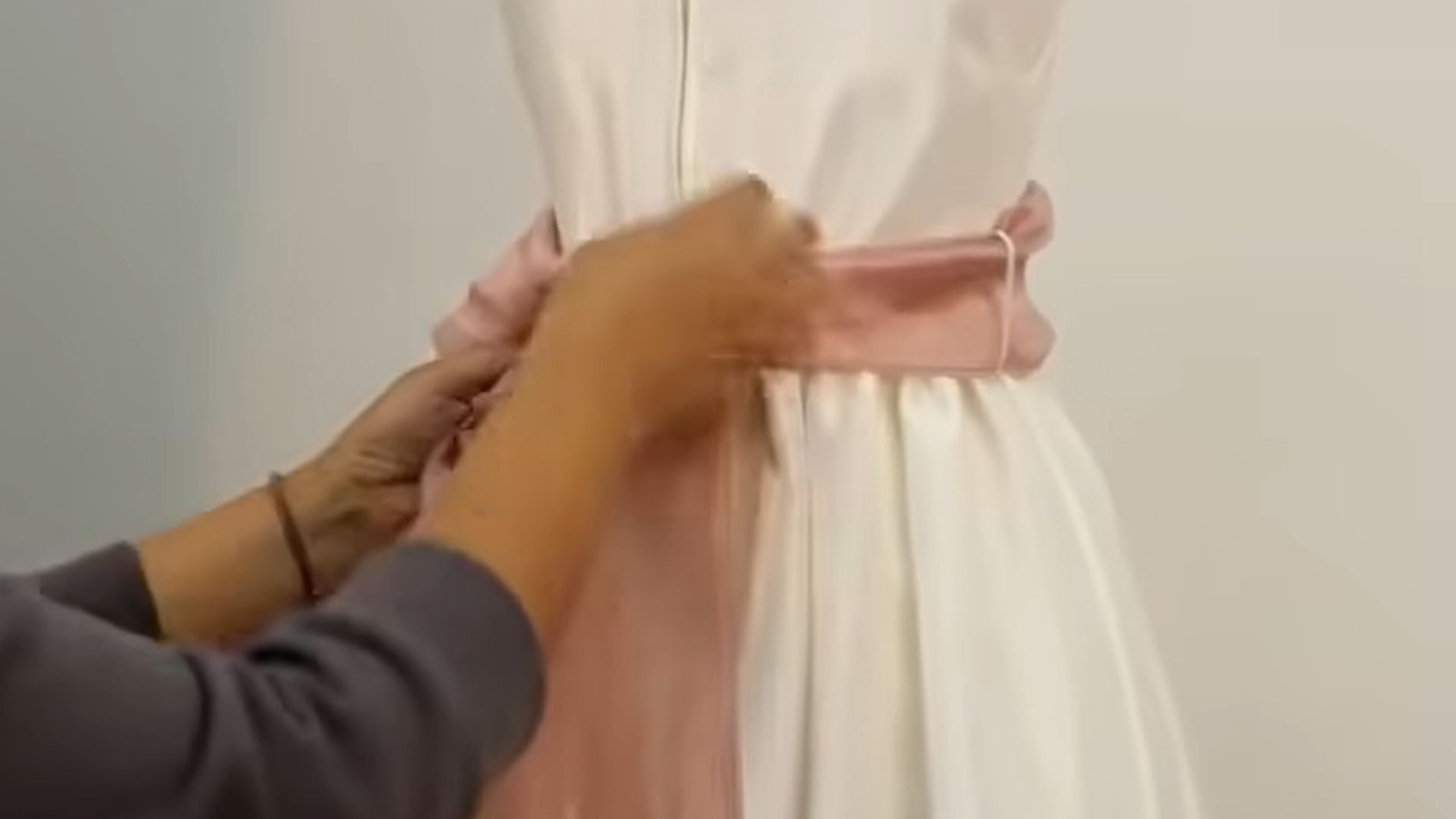"Hands folding and shaping a dusty pink satin sash into a bow at the back of a white dress"