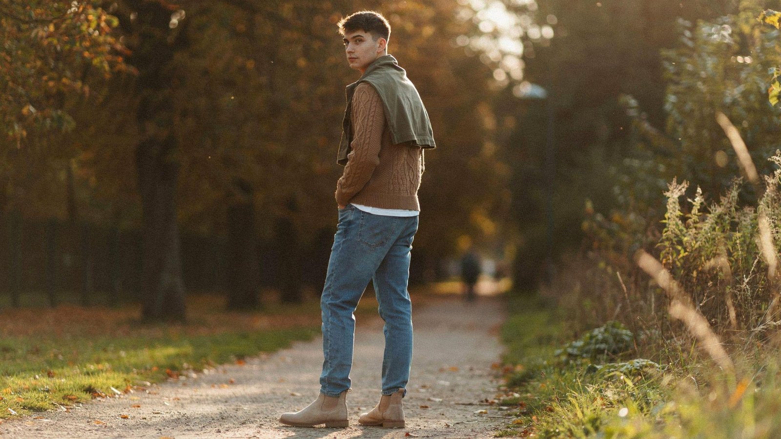 "Young man wearing light blue jeans styled with camel sweater and olive overshirt in a warm earth-tone autumn outfit."