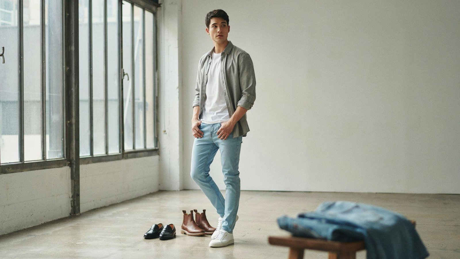 "Fashion editorial of a young man wearing light blue jeans styled with a white t-shirt and grey overshirt in a minimalist natural light studio setting."