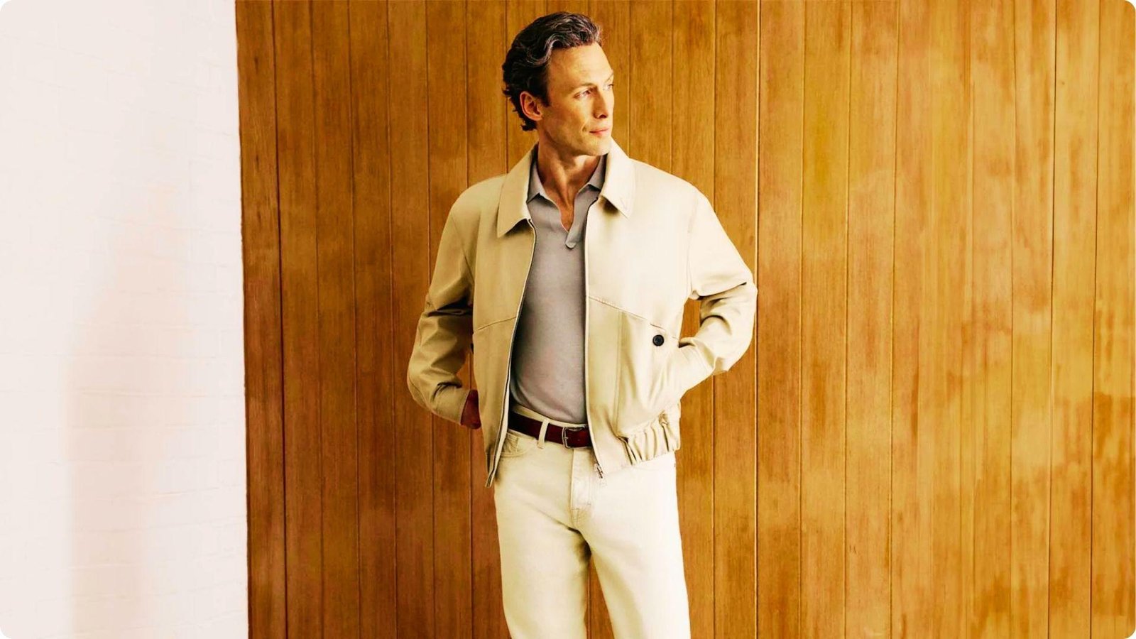 Man in a beige jacket and cream pants against a warm wood-paneled wall.