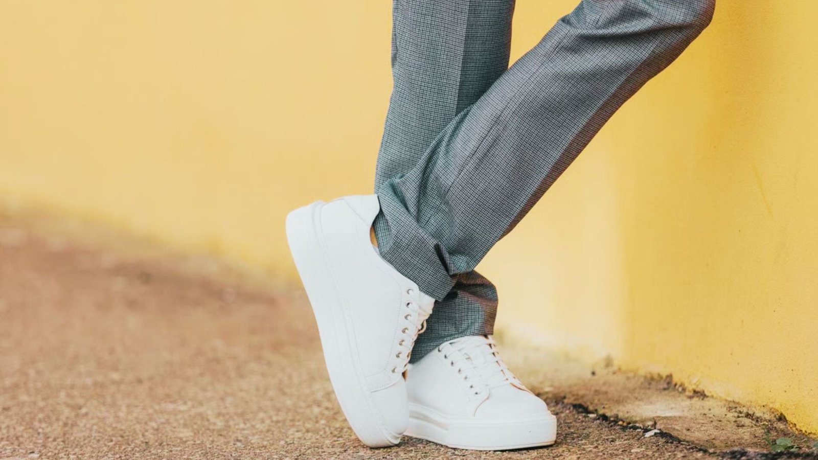 A person wearing white sneakers and pants stands with legs crossed, leaning against a textured orange wall on a concrete surface.