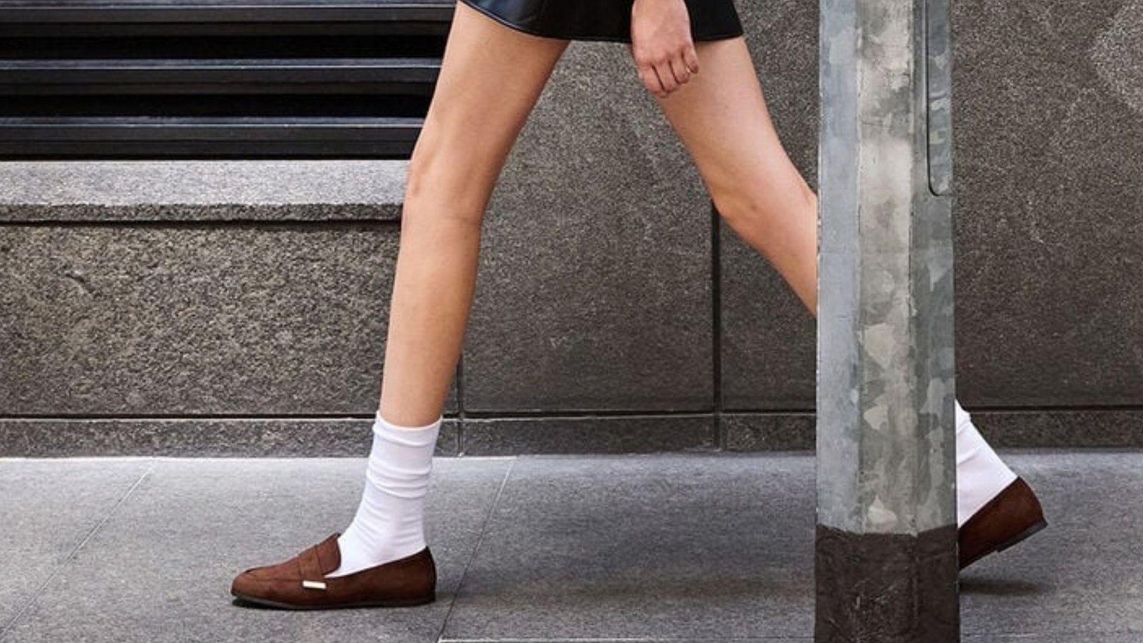 Close-up of a woman's legs wearing brown suede loafers with white socks and a black mini skirt on a city sidewalk.