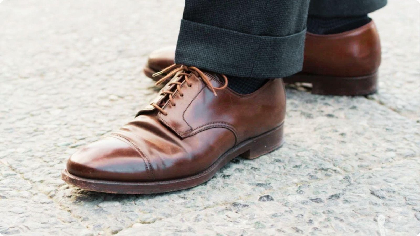 Side view of charcoal dress pants with a neat cuff breaking just above brown leather cap-toe shoes on a concrete surface.
