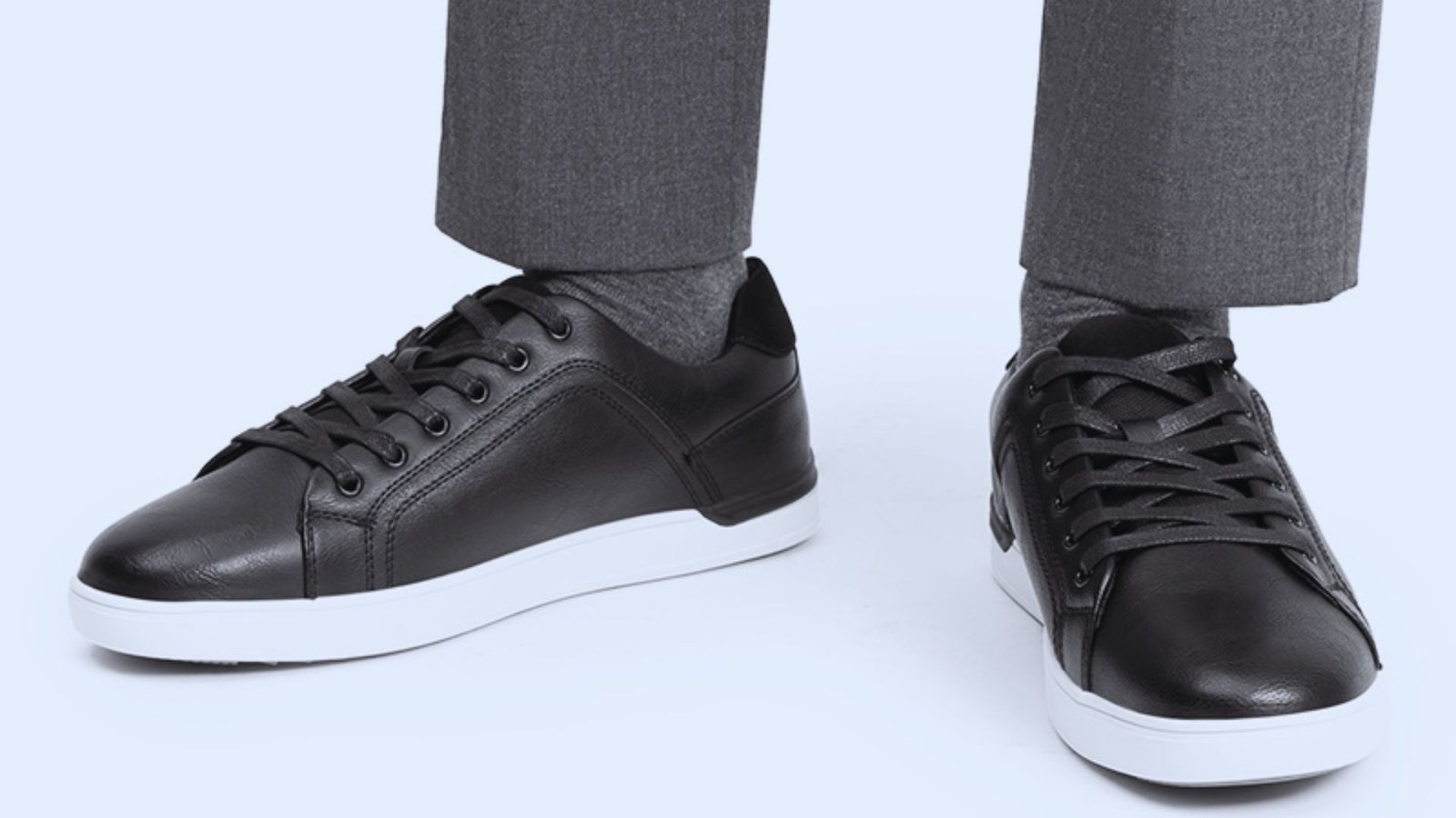 Close-up of black leather sneakers with white soles paired with charcoal dress pants and grey socks on a light blue background