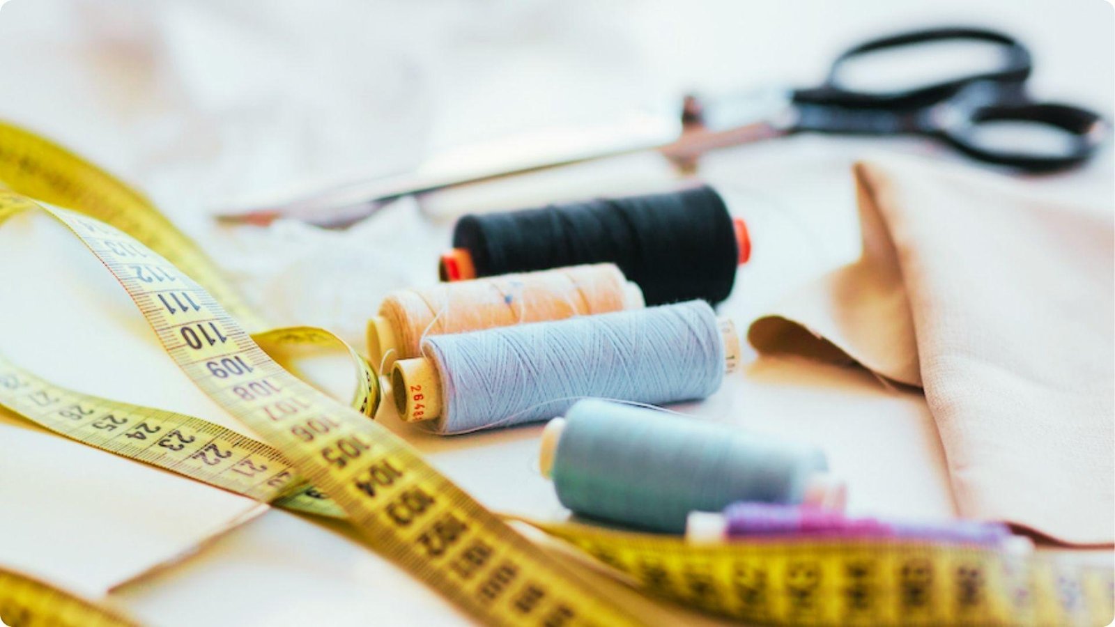 Yellow measuring tape, spools of thread, scissors, and fabric on a tailor’s table.