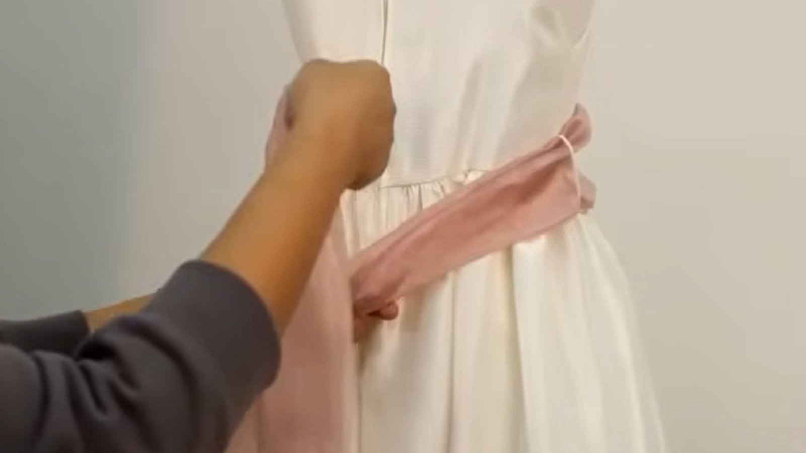 "Hands crossing a dusty pink satin sash at the back of a white dress to begin tying a knot"