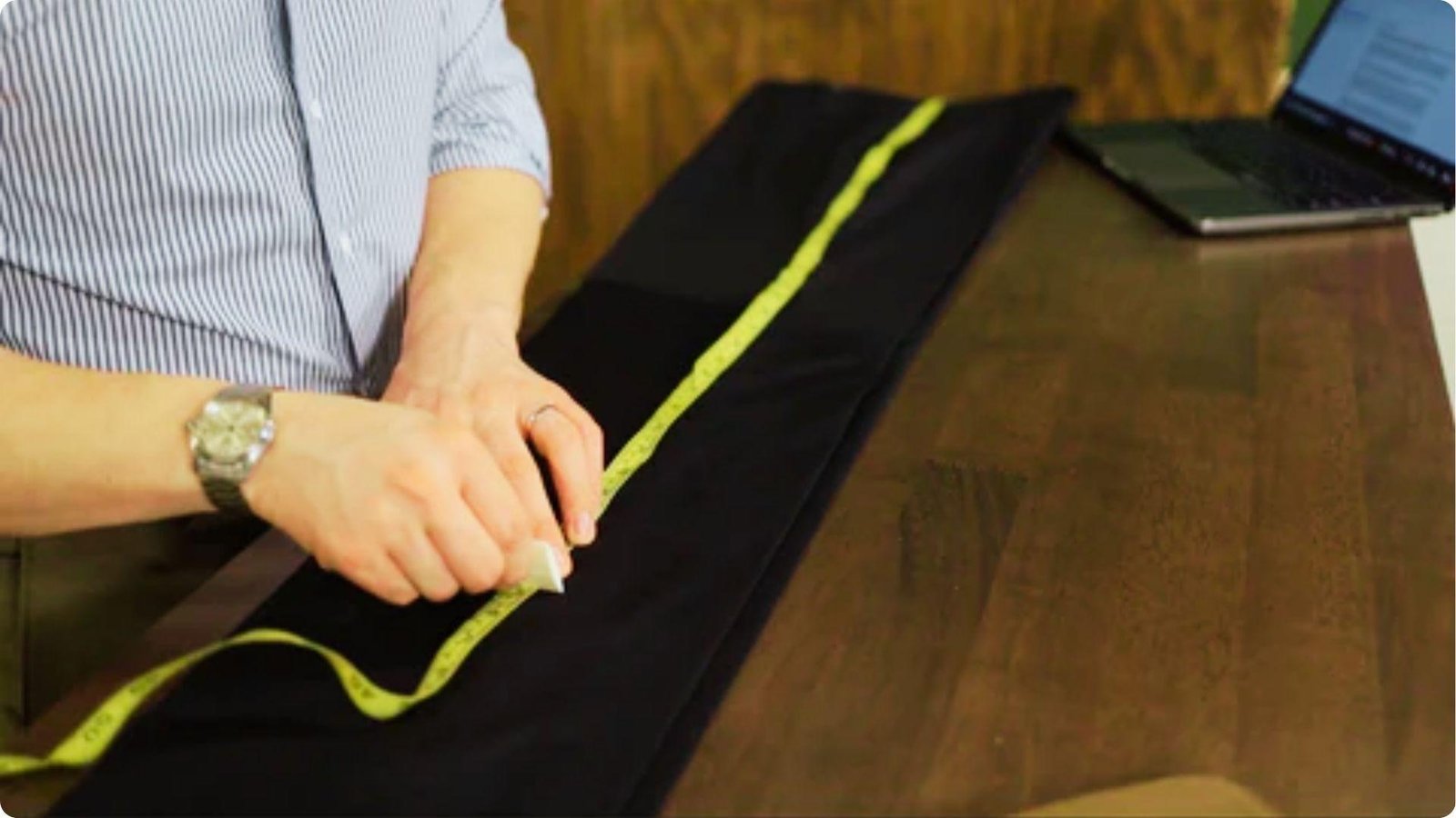 A person in a striped shirt marks a black dress pant leg with chalk while measuring with a yellow tape.