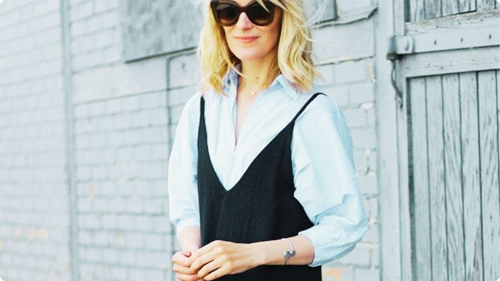 Woman standing against a brick wall in a black slip dress worn over a light blue striped button-down shirt.