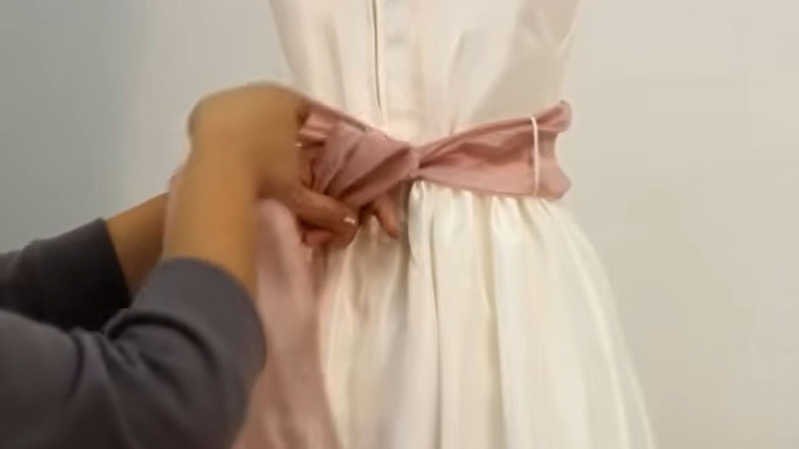 "Hands tightening a dusty pink satin sash around the waist of a white dress"