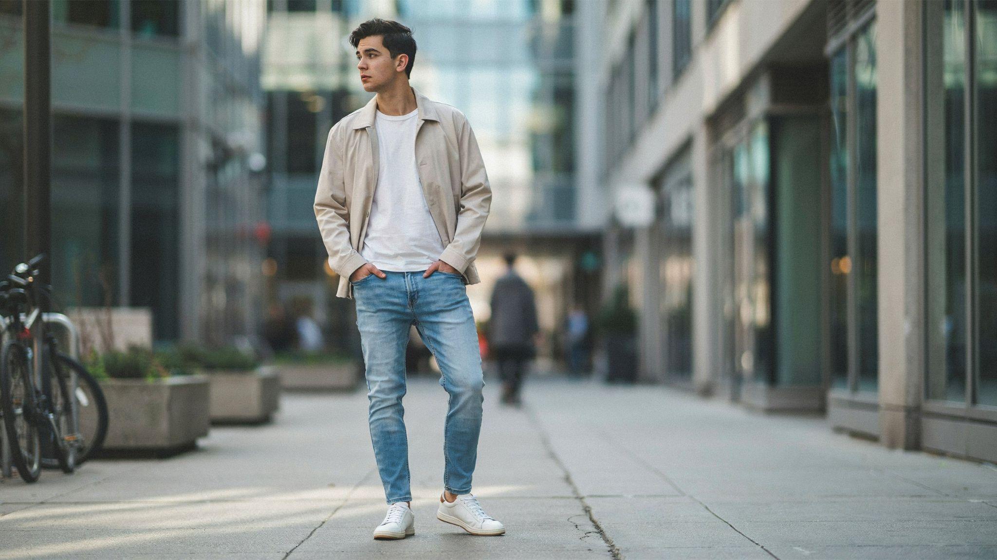 "Modern young man wearing light blue jeans styled with a white t-shirt, beige jacket, and white sneakers in an urban street fashion setting."