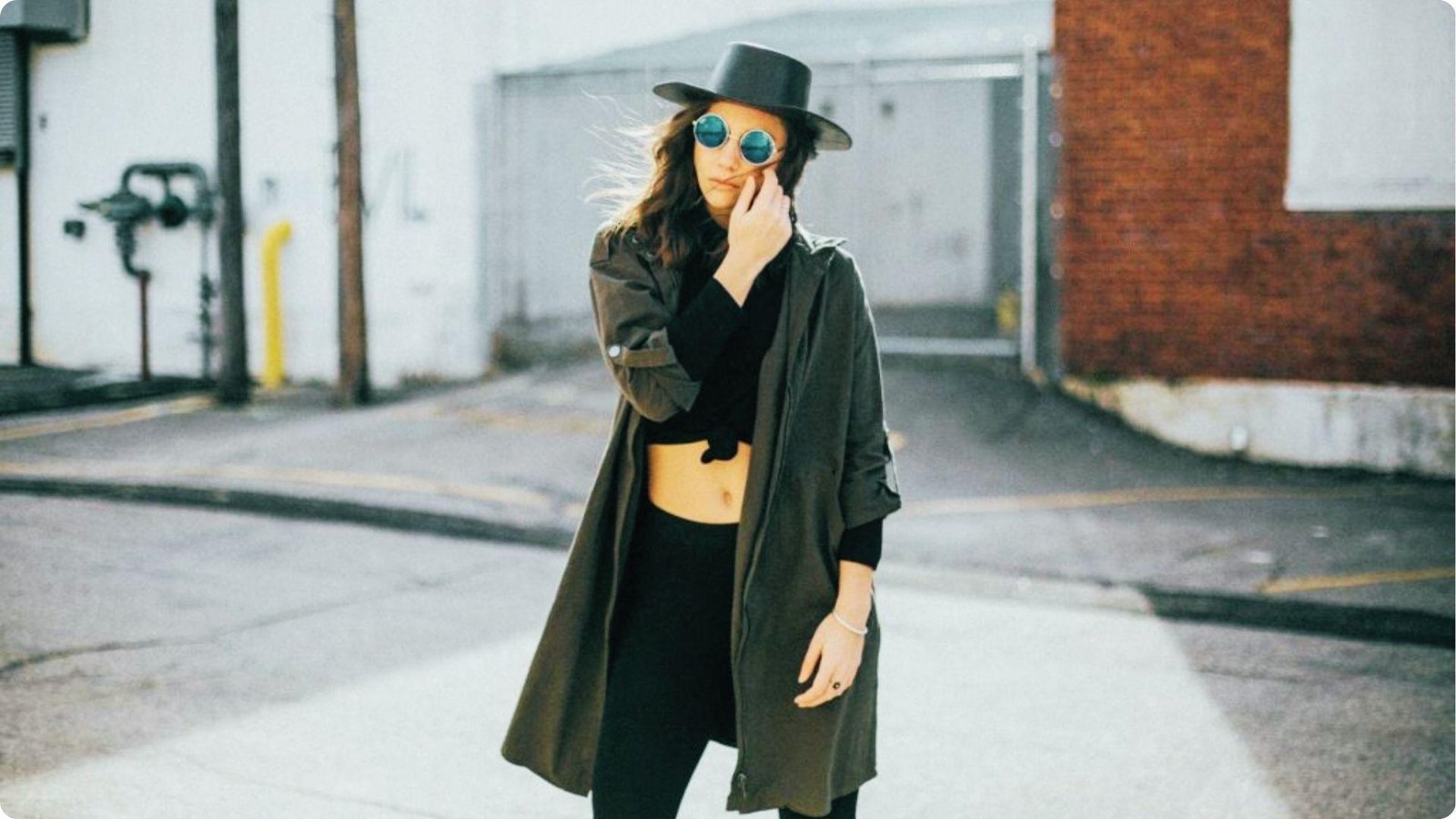 Woman in a long dark coat, black crop top, black pants, and wide-brim hat on a city street.