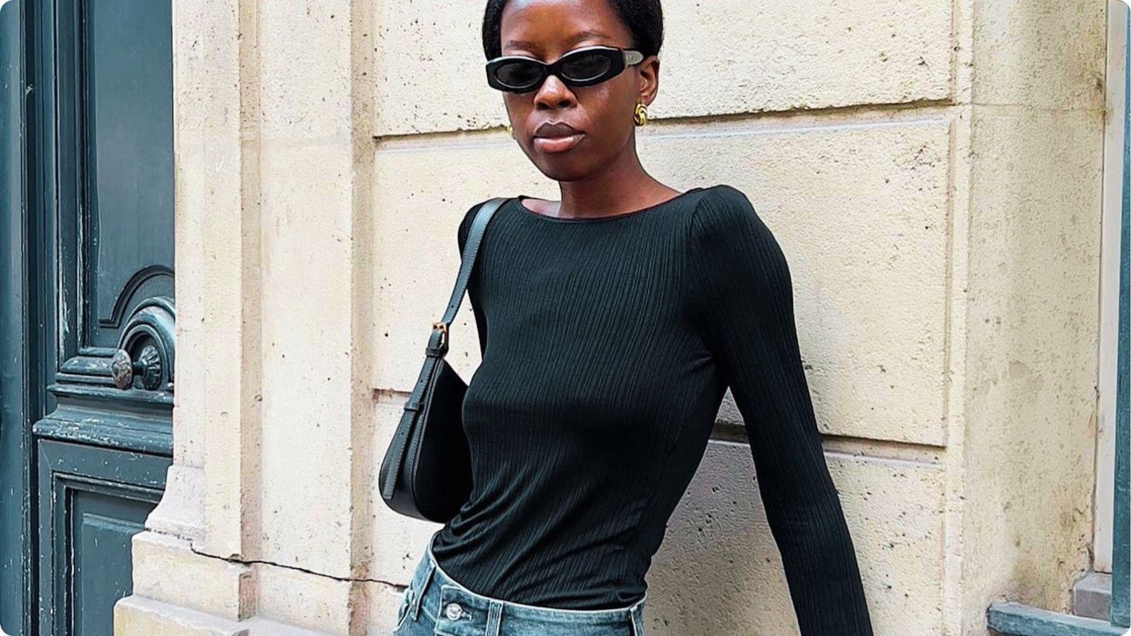 Woman in a fitted black ribbed top and jeans with sunglasses, leaning against a stone wall.