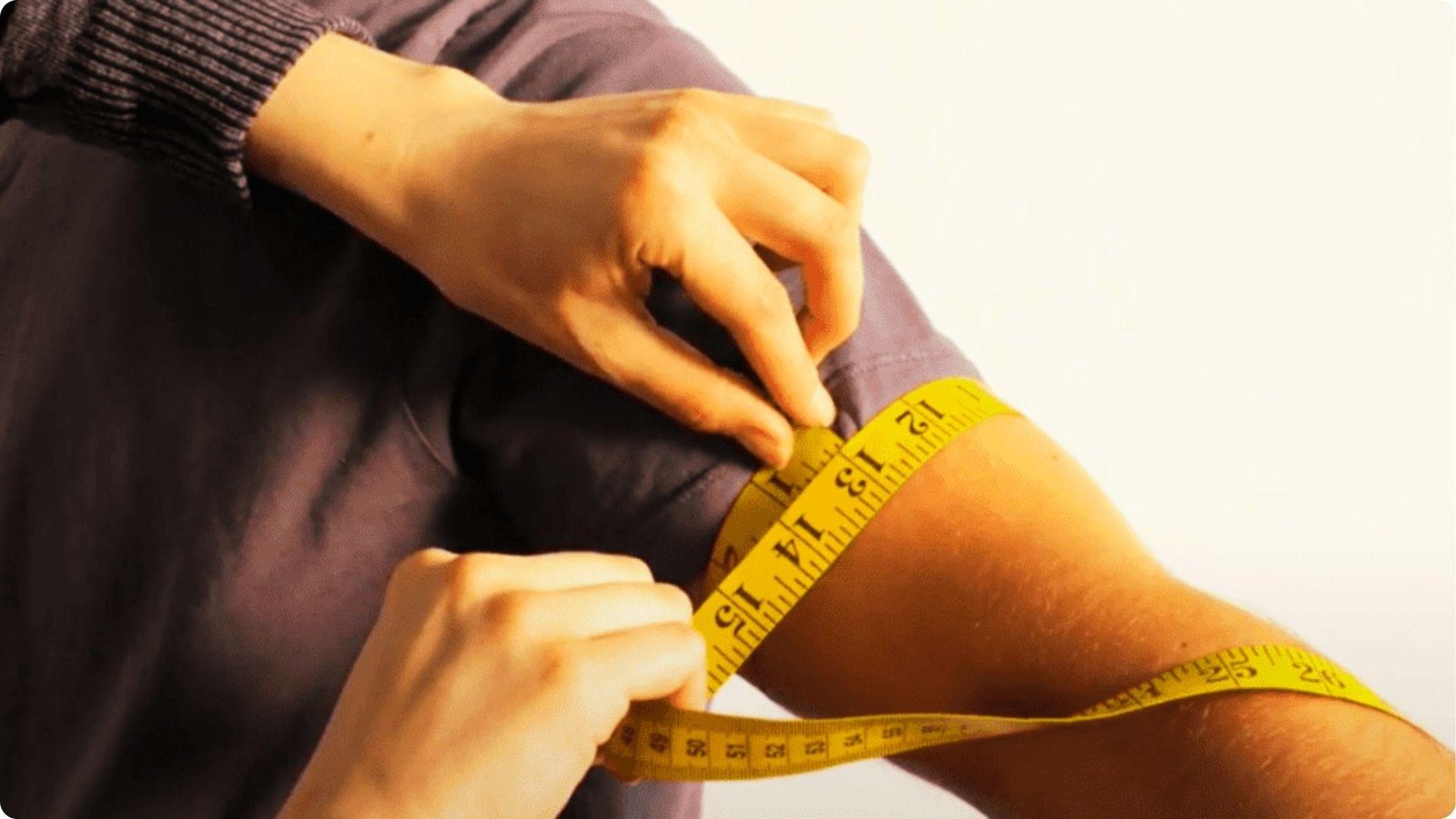 Hands using a yellow measuring tape to measure bicep circumference on a bent arm.