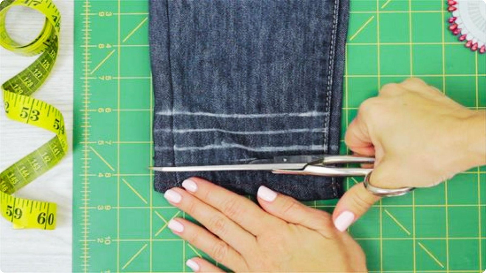 Hands use large scissors to cut excess fabric from a denim pant leg along chalk marks on a green cutting mat.